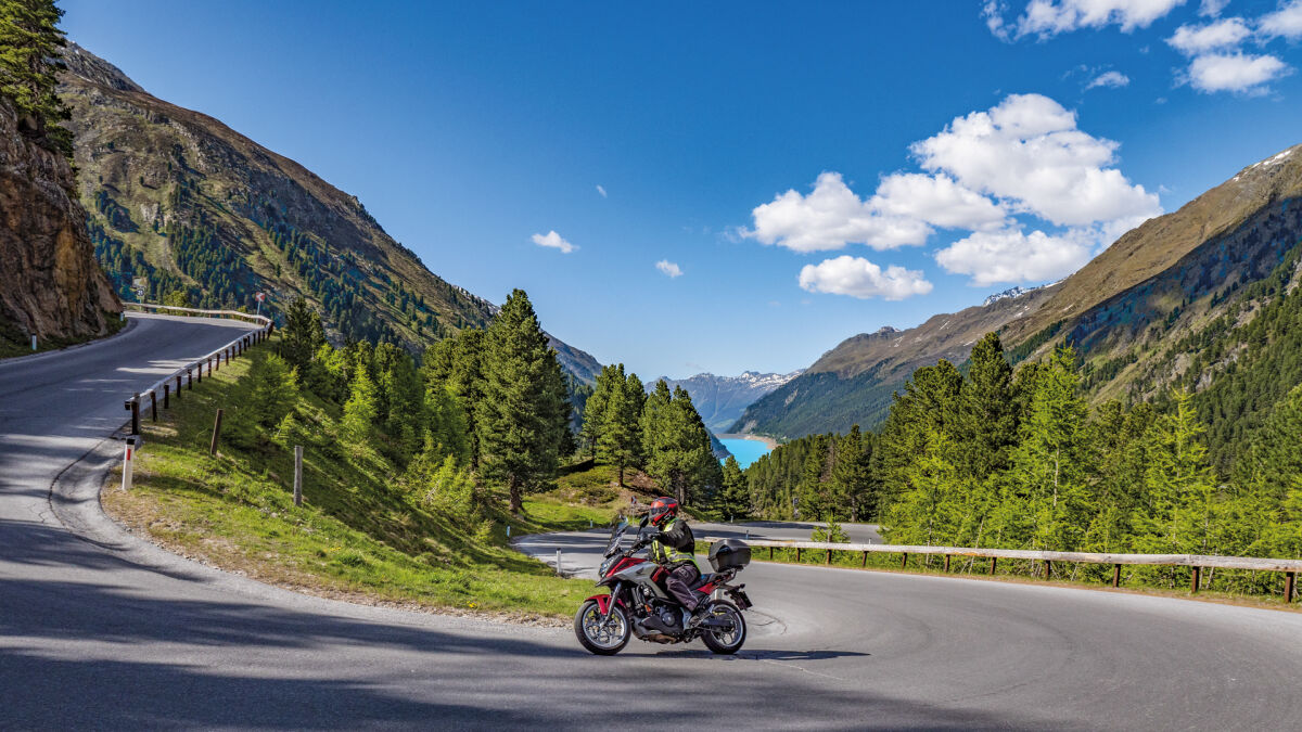 Motorrad in einer Linkskurve, im Tal ein türkisfarbener Stausee, rundum bewaldete Hänge und Gipfel Motorrad in einer Linkskurve, im Tal ein türkisfarbener Stausee, rundum bewaldete Hänge und Gipfel